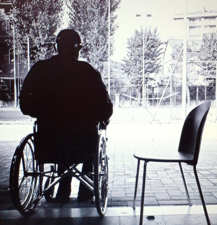 Persona seduta di spalle su una sedia a rotelle, con accanto una sedia vuota, che guarda in direzione di un prato con alcuni alberi intorno sullo sfondo